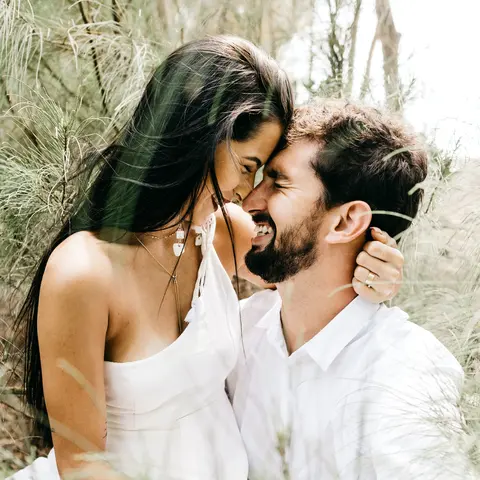 A couple holding each other, forehead to forehead and smiling while standing in a wooded area